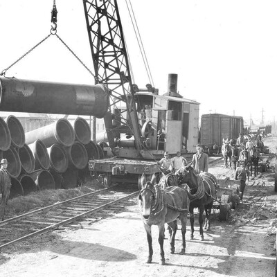 Early 1900's cast iron pipe being offloaded at a rail siding with crane, horses, and wagons.